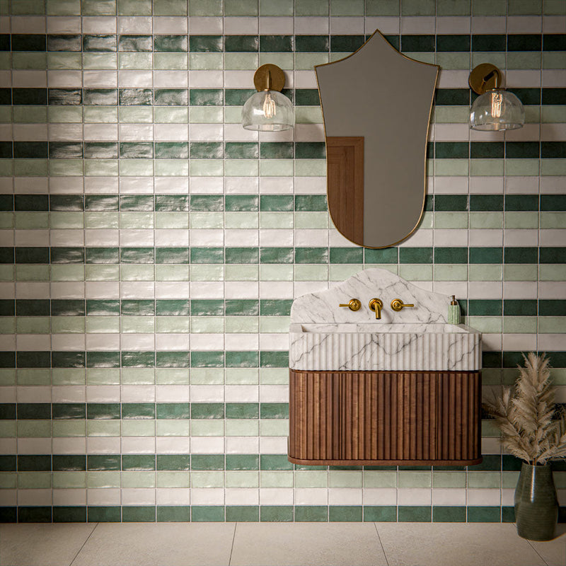 Bathroom interior featuring Cotswold glossy brick wall tiles in mixed green and white tones, installed as a horizontal striped splashback behind a marble vanity unit with brass fixtures and decorative wall lighting.