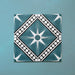 Galata Blue Moroccan Style 20x20cm layout of four tiles featuring a white eight-pointed star motif with black geometric borders and diamond framing on a deep blue background, displayed on a textured teal surface.