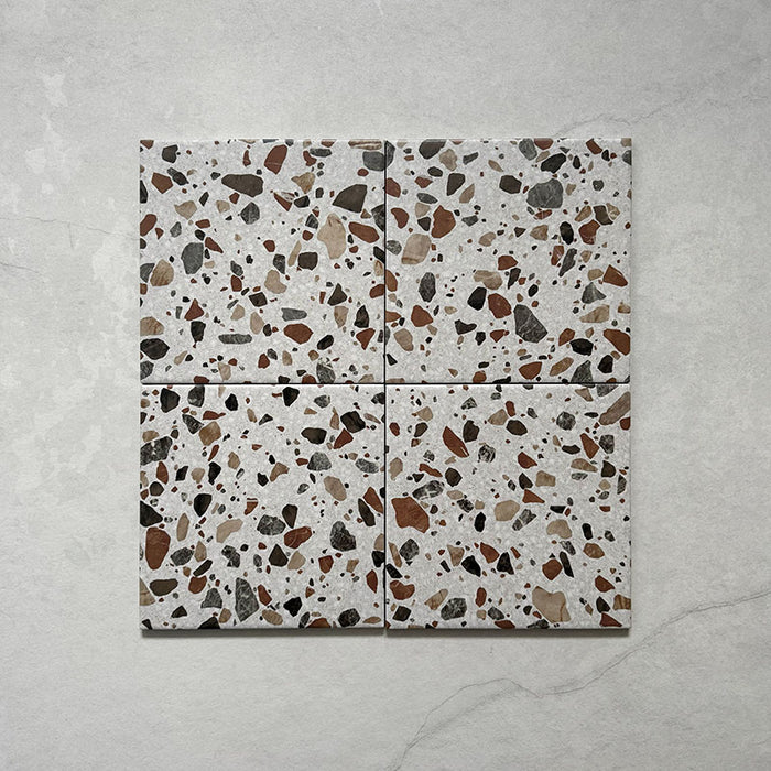 Flatlay featuring four Dino Terrazzo Blanco Drops 20x20cm terrazzo-style ceramic tiles, showing a white base with large stone-effect fragments in earthy brown, terracotta, and grey shades.