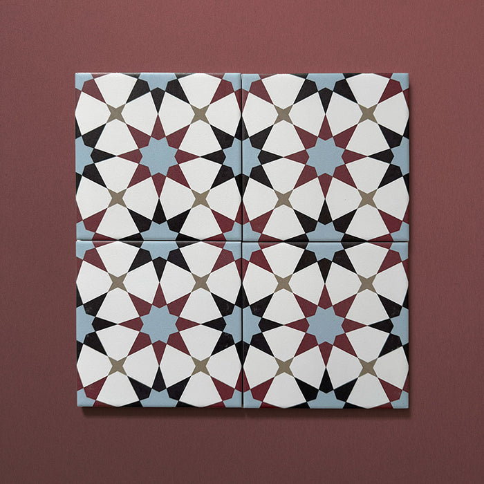 Close-up of four Agraba Mini Atlas 15x15cm Moroccan-style ceramic tiles showing a geometric star pattern in blue, terracotta, black, and white tones on a warm burgundy background.