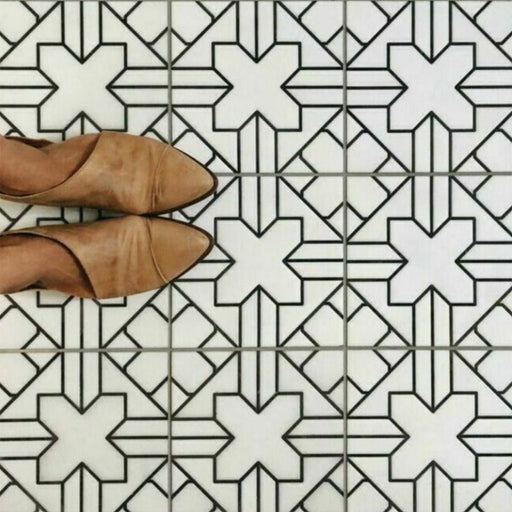 Alhambra Palacio 20x20cm Moroccan tiles with a black geometric cross pattern on a white base, shown from above with tan shoes to highlight the detailed design and matte finish.