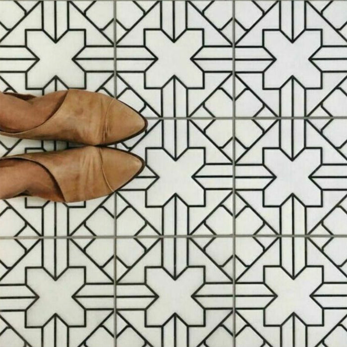 Alhambra Palacio 20x20cm Moroccan tiles with a black geometric cross pattern on a white base, shown from above with tan shoes to highlight the detailed design and matte finish.