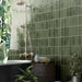 Zellij Botanical Green brick tiles, 6.5cm x 20cm, Moroccan style glazed ceramic wall tiles in rich botanical green tones with natural tonal variation, glossy finish, and handcrafted surface texture, shown in a bathroom setting with a freestanding bath, brass shower fittings, and lush green plants.