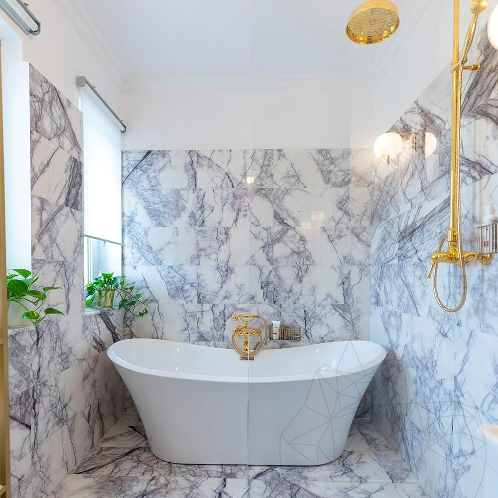 Bathroom interior featuring Fitzrovia Lilac 30.5 x 61cm honed natural marble tiles across walls and floor, showcasing soft lilac veining against a light base, styled with a freestanding bath, brushed brass fittings, and a bright, refined setting.