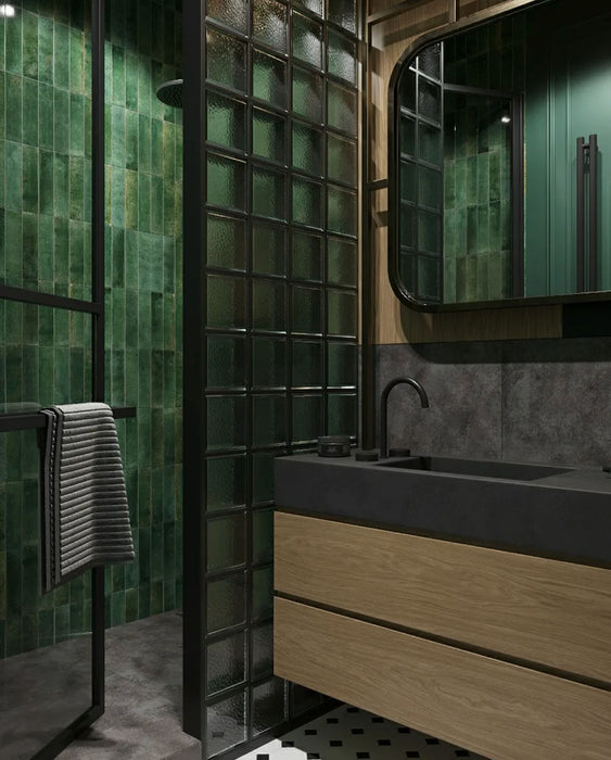 Bathroom setting featuring Mizu Green 6x24cm porcelain wall and floor tiles, showcasing rich tonal variation in deep green hues, paired with glass blocks, dark stone finishes and wood cabinetry for a moody, contemporary, design-led space.