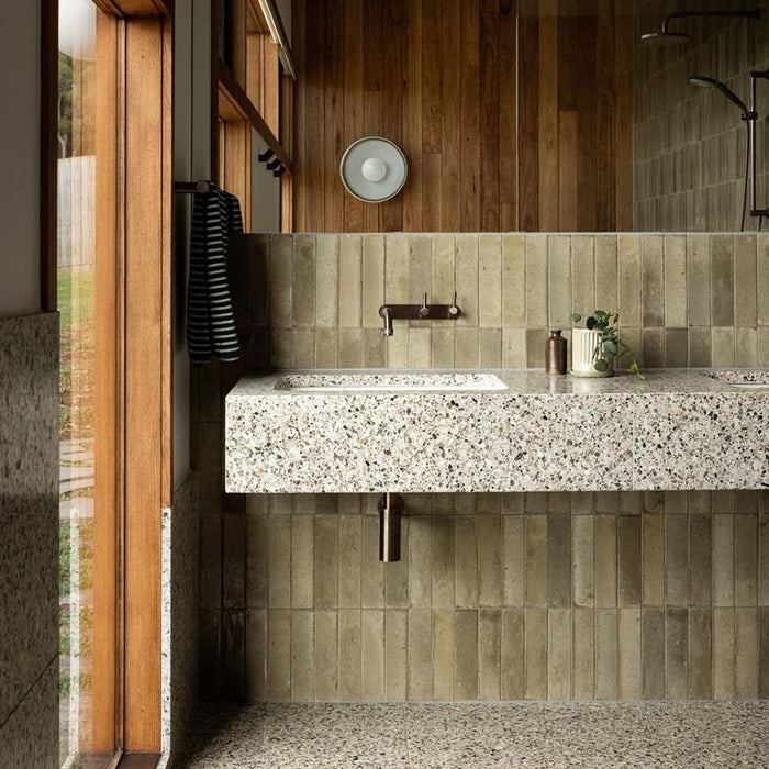 Bathroom featuring Izmir Sage Green gloss brick tiles in a 6 × 25cm ceramic format, installed vertically to create a soft, elongated look, with subtle tonal variation and a high-gloss finish that reflects natural light, paired with warm wood panelling, terrazzo surfaces and minimalist brass fittings for a calm, spa-inspired interior.