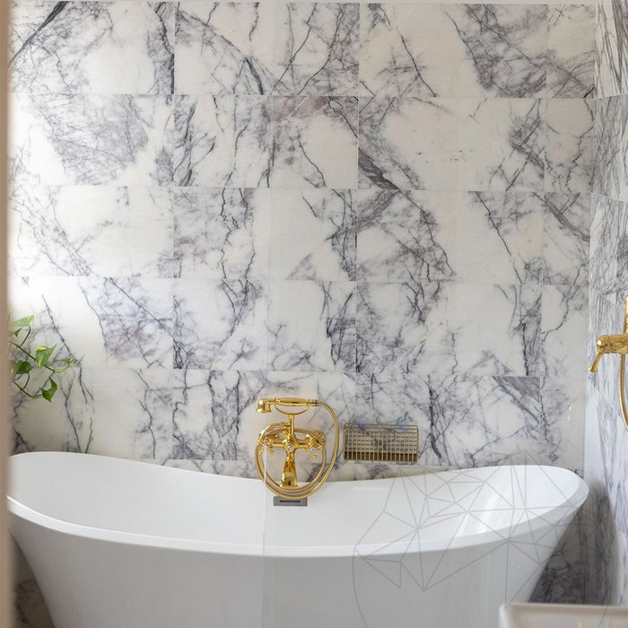 Bathroom interior featuring Fitzrovia Lilac 30.5 x 61cm honed natural marble tiles across walls and floor, showcasing soft lilac veining against a light base, styled with a freestanding bath, brushed brass fittings, and a bright, refined setting.