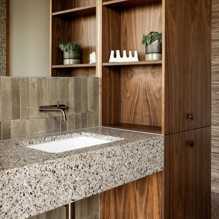 Bathroom featuring Izmir Sage Green gloss brick tiles in a 6 × 25cm ceramic format, installed vertically to create a soft, elongated look, with subtle tonal variation and a high-gloss finish that reflects natural light, paired with warm wood panelling, terrazzo surfaces and minimalist brass fittings for a calm, spa-inspired interior.