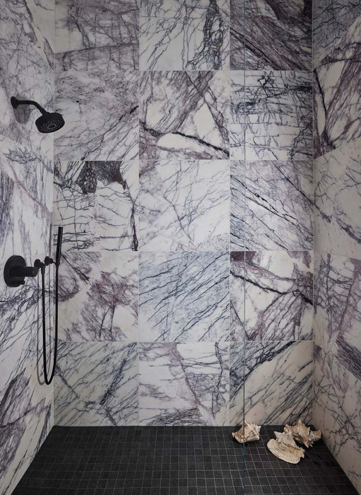 Shower interior featuring Fitzrovia Lilac 30.5 x 61cm honed natural marble tiles across the walls, showcasing striking lilac and charcoal veining against a soft white base, paired with matte black brassware and a contrasting dark mosaic floor for a bold, design-led finish.