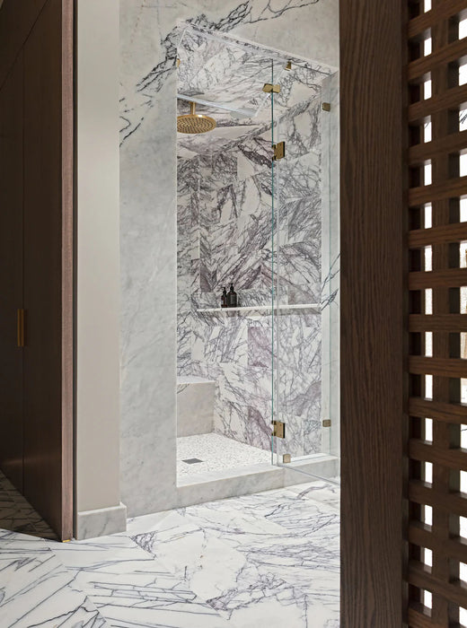 Luxury bathroom interior featuring Fitzrovia Lilac 30.5 x 61cm honed natural marble tiles across walls and floor, showcasing expressive lilac and charcoal veining, styled with a walk-in shower, brushed brass fittings, and a refined, spa-like setting.