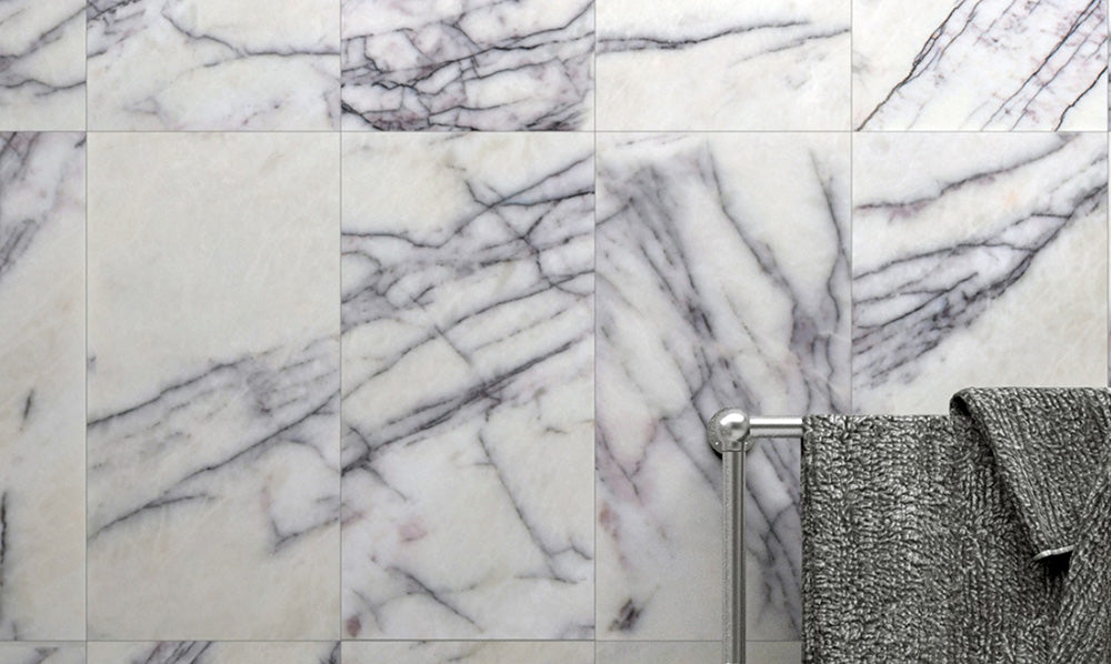 Bathroom wall detail featuring Fitzrovia Lilac 30.5 x 61cm honed natural marble tiles, showcasing soft white tones with flowing lilac-grey veining, styled with a chrome towel rail and textured grey towel for a calm, contemporary finish.