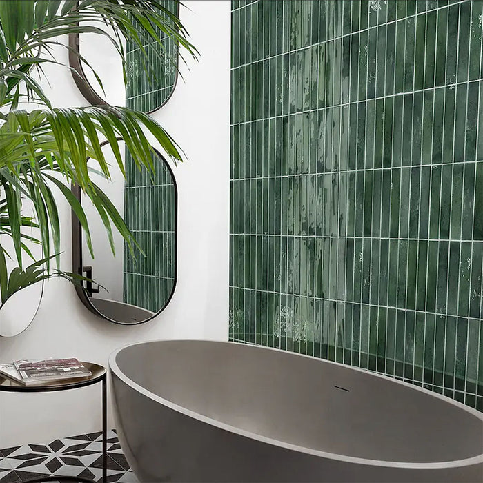 Modern bathroom featuring Izmir Brick Green Gloss 6x25cm ceramic wall tiles, styled with a freestanding grey bath, geometric floor tiles, curved mirrors, and lush green plants for a fresh, contemporary look.