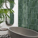 Modern bathroom featuring Izmir Brick Green Gloss 6x25cm ceramic wall tiles, styled with a freestanding grey bath, geometric floor tiles, curved mirrors, and lush green plants for a fresh, contemporary look.
