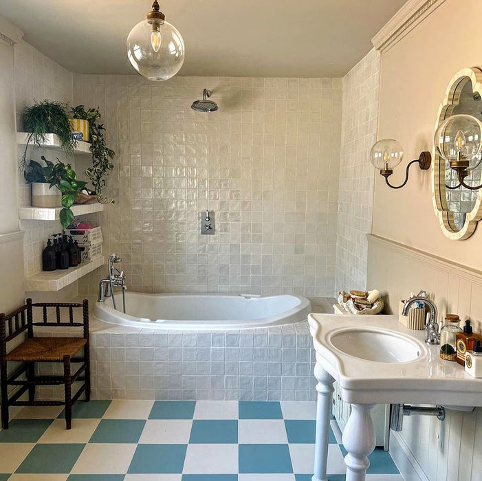 Bathroom setting featuring Zellij Oyster Square Moroccan-style tiles in a 10cm x 10cm format, with a glossy off-white glaze and subtle handmade texture, used on walls around a freestanding bath and paired with a classic pedestal sink and patterned floor tiles.