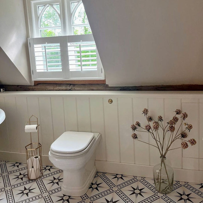 “Galata White Moroccan Style 20x20cm tiles in a bright bathroom featuring white panelled walls, a traditional toilet, and patterned floor tiles with black star and diamond motifs, complemented by a gold toilet roll holder and vase with dried flowers.