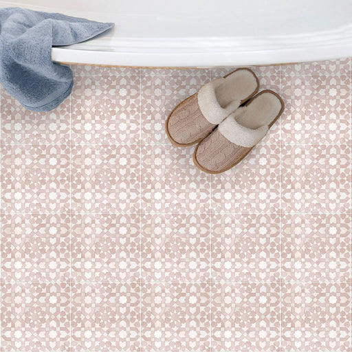 Bedouin Tetuan Rose Moroccan-style 15x15cm tiles used as bathroom flooring beneath a white bathtub. The tiles feature a soft blush pink and white geometric pattern with a handcrafted matte texture, paired with beige slippers and a blue towel for a cosy, spa-like aesthetic.