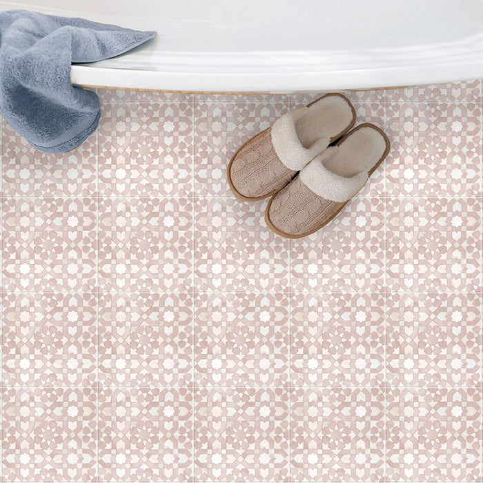 Bedouin Tetuan Rose Moroccan-style 15x15cm tiles used as bathroom flooring beneath a white bathtub. The tiles feature a soft blush pink and white geometric pattern with a handcrafted matte texture, paired with beige slippers and a blue towel for a cosy, spa-like aesthetic.