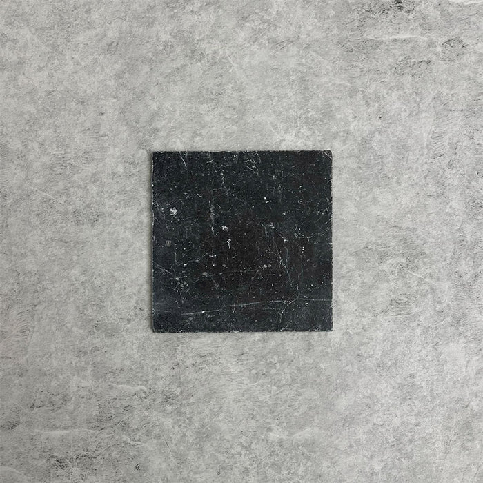 Single Fitzrovia Black 15.2x15.2cm tumbled natural marble tile displayed on a neutral grey surface, showcasing deep charcoal tones, subtle natural veining, and a softly aged, textured finish.