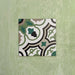 Single Europa Budapest 20x20cm Victorian Moroccan style tile displayed against a textured green wall, showcasing an intricate geometric pattern in green, cream, black and taupe tones in a close-up product setting.