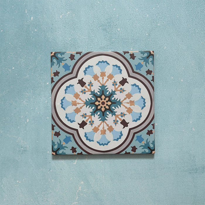 Cabana Bayamo Moroccan style patterned tile in a 20cm x 20cm format, featuring an intricate geometric floral design in soft blue, cream, teal and warm brown tones, shown against a textured blue background.