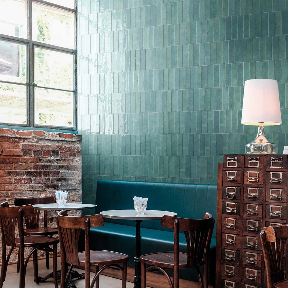Madison Aquamarine Brick Wall Tiles 6x24.6cm — shown here in a stylish café setting, these glossy teal-green tiles bring depth and vibrancy to the space. Their handcrafted appearance and reflective surface create a lively, modern look that pairs beautifully with natural wood and industrial details.