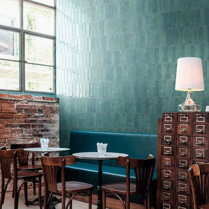 Madison Aquamarine Brick Wall Tiles 6x24.6cm — shown here in a stylish café setting, these glossy teal-green tiles bring depth and vibrancy to the space. Their handcrafted appearance and reflective surface create a lively, modern look that pairs beautifully with natural wood and industrial details.