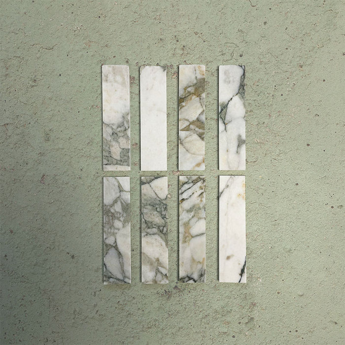 Eight Fitzrovia Calacatta Green 5 x 20cm honed natural marble tiles arranged on a soft green backdrop, showcasing a light base with delicate grey and green veining, accented by subtle warm undertones that highlight the natural variation of the stone.