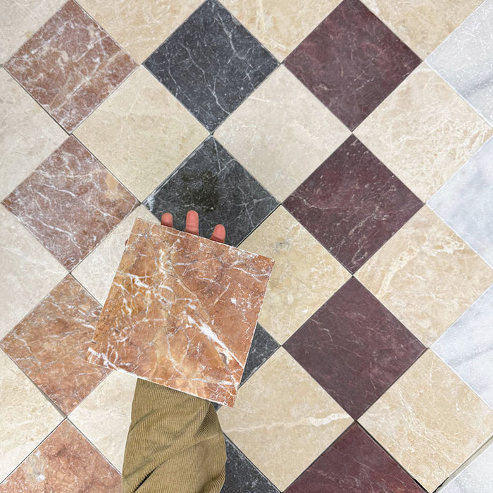 Hand holding Fitzrovia Rosso 15.2x15.2cm tumbled natural marble tile against a multi-tone chequerboard floor, showcasing warm terracotta hues, natural veining, and varied marble colours in a classic patterned layout.
