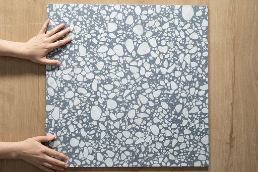 Close-up of Chunks POP Terrazzo Blue 60 × 60cm terrazzo-effect porcelain tiles, showcasing a soft blue base with scattered white aggregate detailing, photographed against natural wood flooring for scale and texture reference.