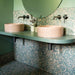 Contemporary bathroom featuring Chunks POP Terrazzo Green 60 × 60cm terrazzo-effect porcelain wall and floor tiles, styled with blush pink basins, sage green surfaces, round mirrors, and dark brushed metal brassware.