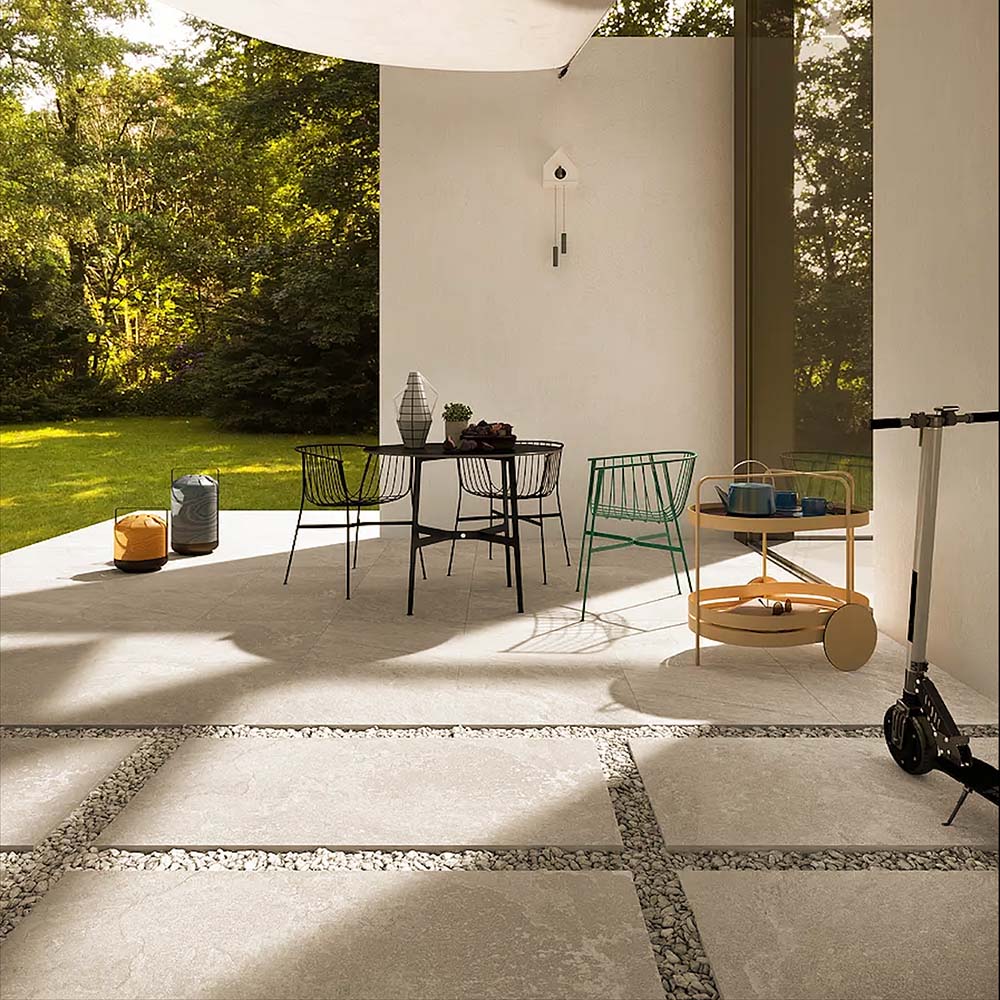 Outdoor patio featuring Cocoon Stone Light 2cm 60 × 90cm porcelain tiles, laid in a modern grid with gravel joints. Soft light stone effect tones create a bright, airy feel, complemented by contemporary furniture and surrounding greenery for a relaxed, indoor-outdoor setting.