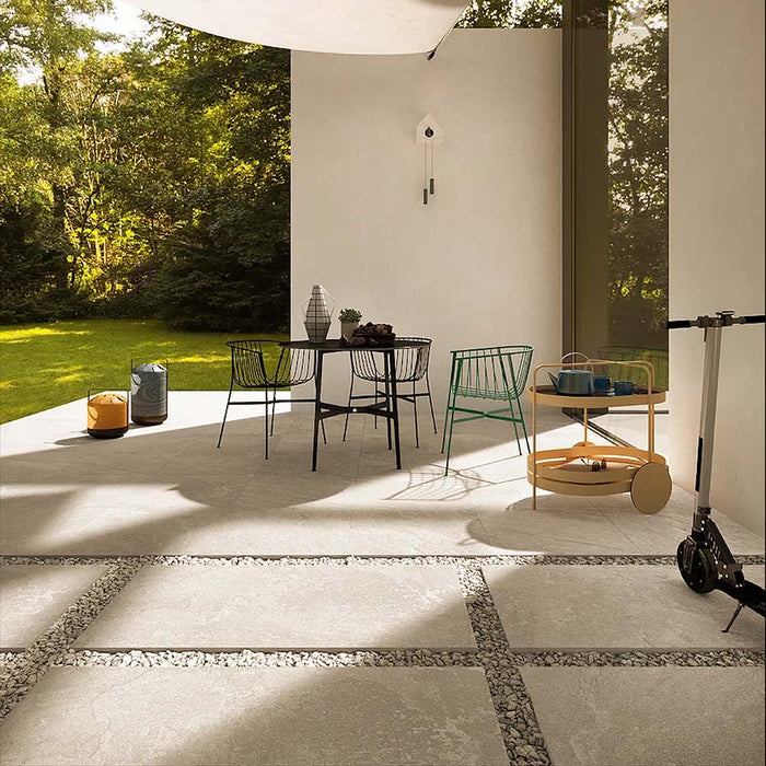 Outdoor patio featuring Cocoon Stone Light 2cm 60 × 90cm porcelain tiles, laid in a modern grid with gravel joints. Soft light stone effect tones create a bright, airy feel, complemented by contemporary furniture and surrounding greenery for a relaxed, indoor-outdoor setting.