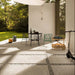 Outdoor patio featuring Cocoon Stone Light 2cm 60 × 90cm porcelain tiles, laid in a modern grid with gravel joints. Soft light stone effect tones create a bright, airy feel, complemented by contemporary furniture and surrounding greenery for a relaxed, indoor-outdoor setting.