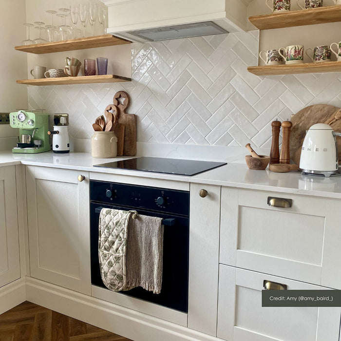 Kitchen splashback featuring Zellij White Gloss Brick Moroccan-style tiles in a 6.5cm x 20cm format, laid in a herringbone pattern, with a glossy white glaze and subtle handmade texture, styled with shaker cabinetry, open wooden shelving, and brass accents.