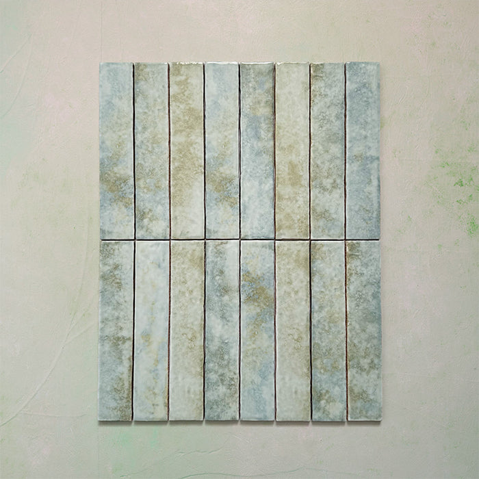 Product display of Elazar Agave Green 5x25cm glossy wall tiles in soft sage and green tones, featuring handcrafted edges and subtle tonal variation against a textured plaster-style backdrop.