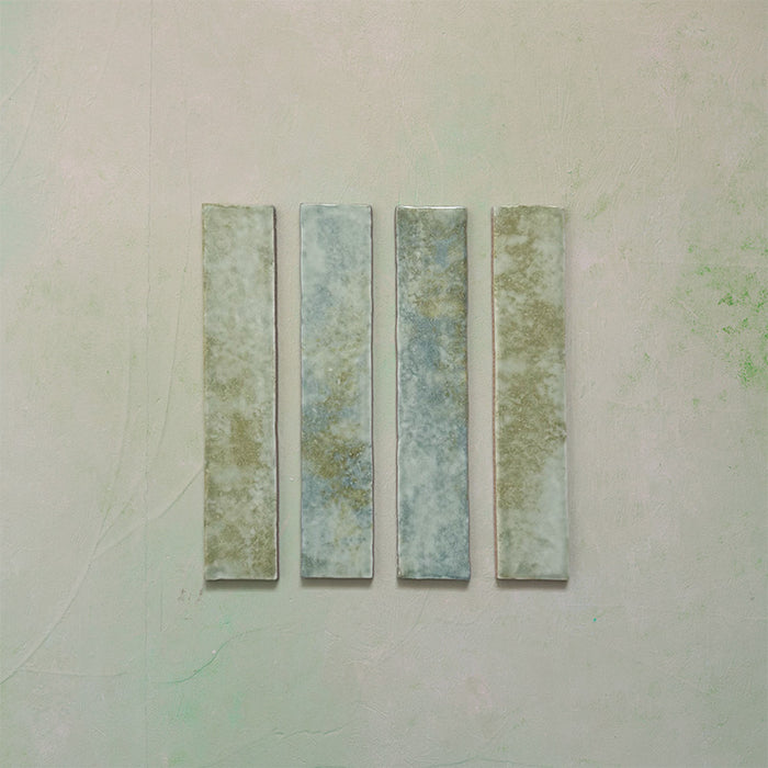 Product display of Elazar Agave Green 5x25cm glossy wall tiles in soft sage and green tones, featuring handcrafted edges and subtle tonal variation against a textured plaster-style backdrop.