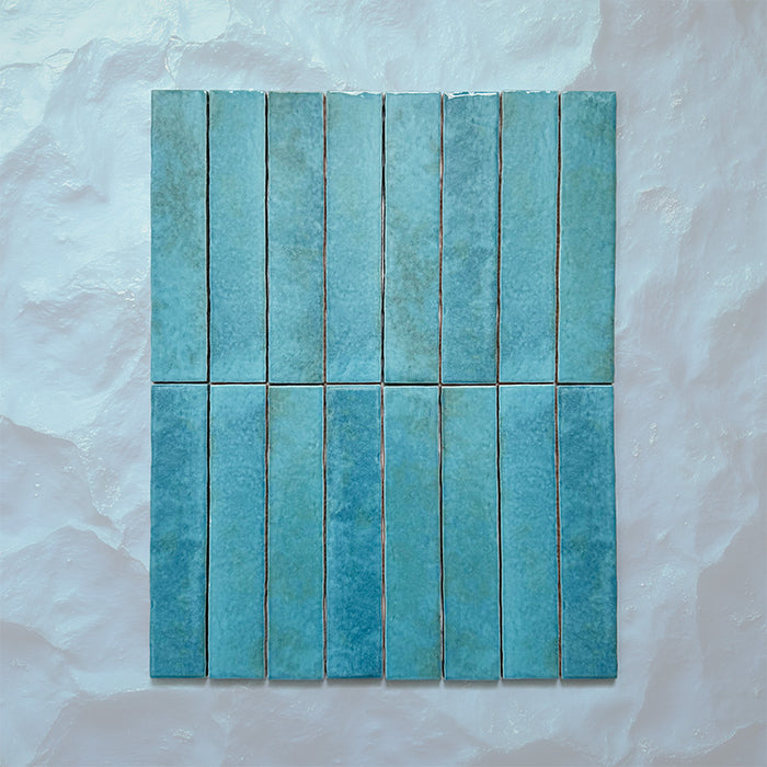 Product display of Elazar Azul Blue 5x25cm glossy wall tiles in vibrant blue tones, featuring handcrafted edges and subtle tonal variation against a textured plaster-style backdrop.