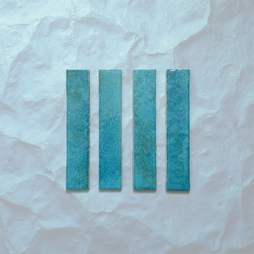 Product display of Elazar Azul Blue 5x25cm glossy wall tiles in vibrant blue tones, featuring handcrafted edges and subtle tonal variation against a textured plaster-style backdrop.