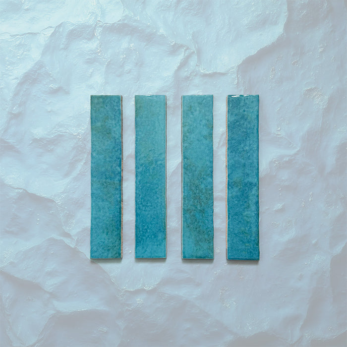 Product display of Elazar Azul Blue 5x25cm glossy wall tiles in vibrant blue tones, featuring handcrafted edges and subtle tonal variation against a textured plaster-style backdrop.