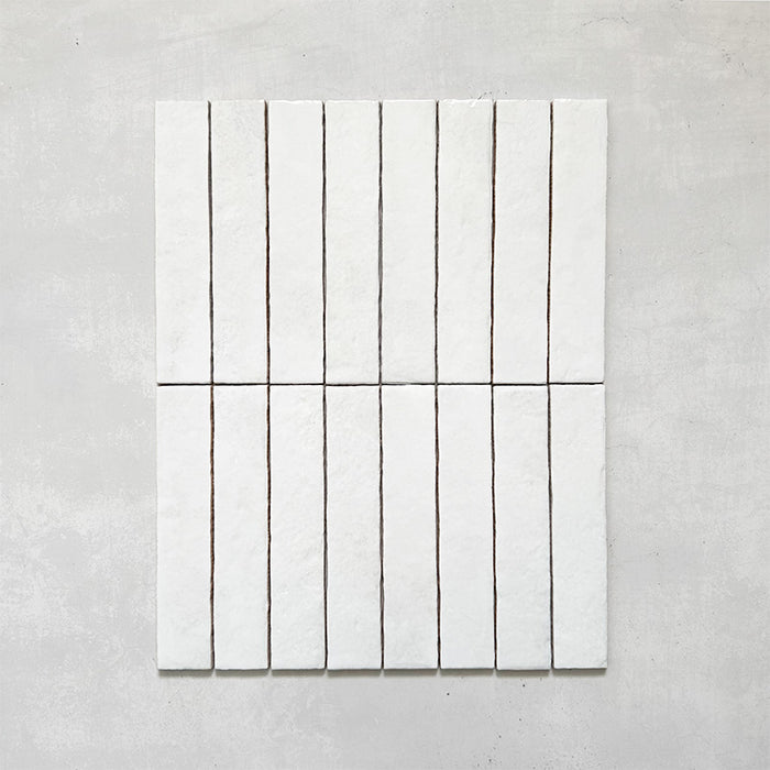 Product display of Elazar Blanco 5x25cm glossy wall tiles in soft white tones, featuring handcrafted edges and subtle surface variation against a textured plaster-style backdrop.