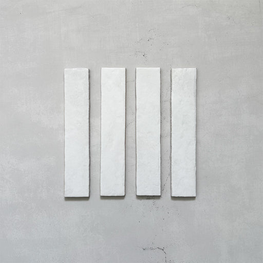 Product display of Elazar Blanco 5x25cm glossy wall tiles in soft white tones, featuring handcrafted edges and subtle surface variation against a textured plaster-style backdrop.