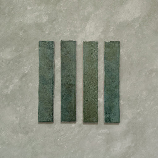 Product display of Elazar Forest Green 5x25cm glossy wall tiles in deep forest green tones, featuring handcrafted edges and rich tonal variation against a textured plaster-style backdrop.