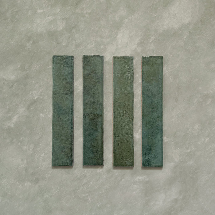 Product display of Elazar Forest Green 5x25cm glossy wall tiles in deep forest green tones, featuring handcrafted edges and rich tonal variation against a textured plaster-style backdrop.