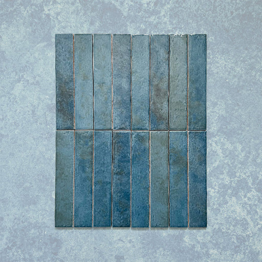 Product display of Elazar Prussian 5x25cm glossy wall tiles in deep Prussian blue tones, featuring handcrafted edges and rich tonal variation against a textured plaster-style backdrop.
