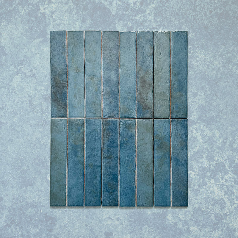 Product display of Elazar Prussian 5x25cm glossy wall tiles in deep Prussian blue tones, featuring handcrafted edges and rich tonal variation against a textured plaster-style backdrop.