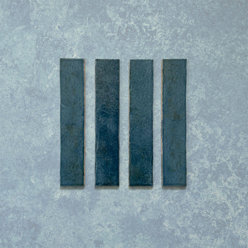 Product display of Elazar Prussian 5x25cm glossy wall tiles in deep Prussian blue tones, featuring handcrafted edges and rich tonal variation against a textured plaster-style backdrop.