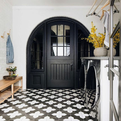 Alhambra Cathedral 20x20cm Moroccan tiles used in a modern entryway with black double doors, wooden bench, and yellow floral accents — featuring a bold black and white cross pattern ideal for hallways or utility spaces.