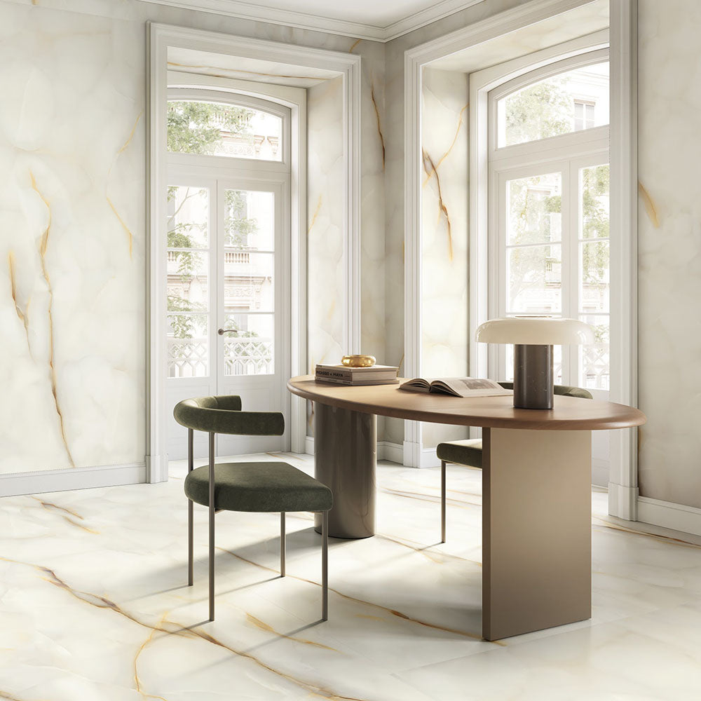 Dining space featuring Fantasia Pearl Onyx Silk Matt 60 × 120cm stone effect floor and wall tiles, with soft cream tones and delicate gold veining, paired with a wooden table, upholstered chairs, and large arched windows.