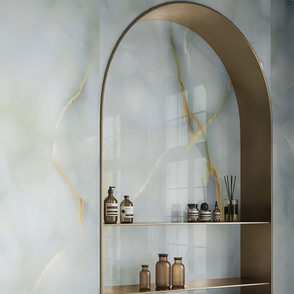 Bathroom interior featuring Fantasia Turchese Blue Onyx Polished 60 × 120cm stone effect wall tiles, showcasing soft blue-grey tones with striking gold veining, paired with an arched metallic shelving niche and warm-toned accessories for a refined, contemporary finish.