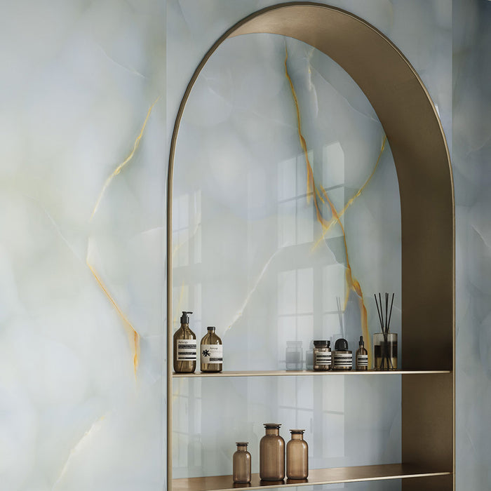 Bathroom interior featuring Fantasia Turchese Blue Onyx Polished 60 × 120cm stone effect wall tiles, showcasing soft blue-grey tones with striking gold veining, paired with an arched metallic shelving niche and warm-toned accessories for a refined, contemporary finish.